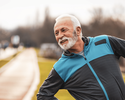 Older man with beard in blue jacket on path doing some stretching.